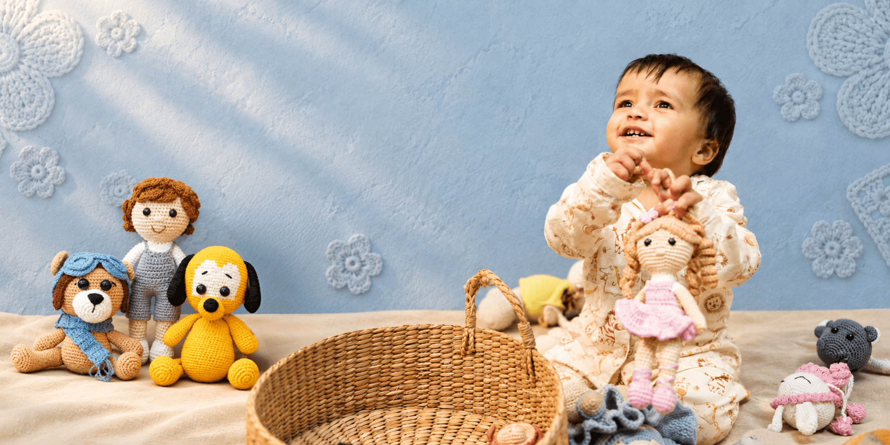 Baby sitting with handmade crochet toys, including a pilot dog, a boy doll, a yellow dog, and a girl doll in a pink dress